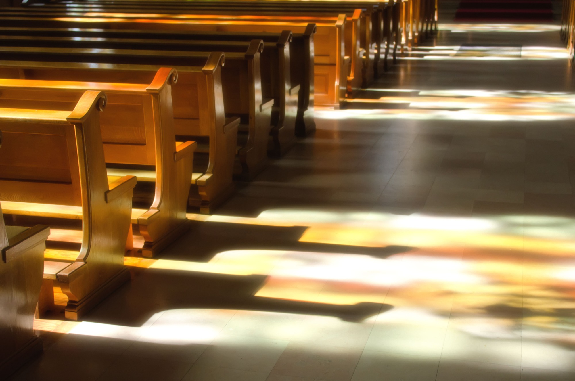 Sunlight streams through stained glass, casting colorful shadows on polished wooden pews in a serene church interior.