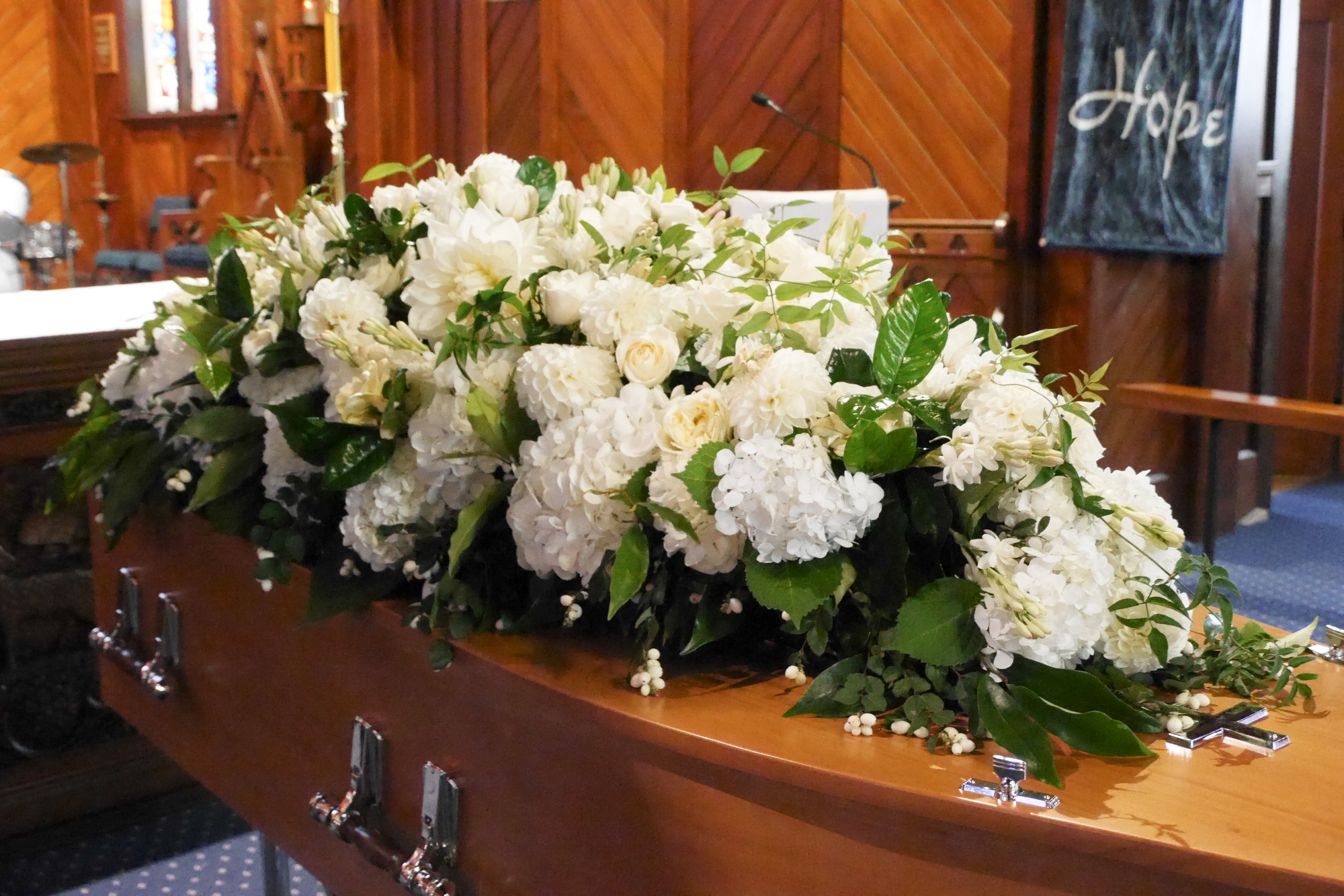 A beautifully arranged display of white flowers atop a wooden casket in a somber setting, symbolizing remembrance and mourning.