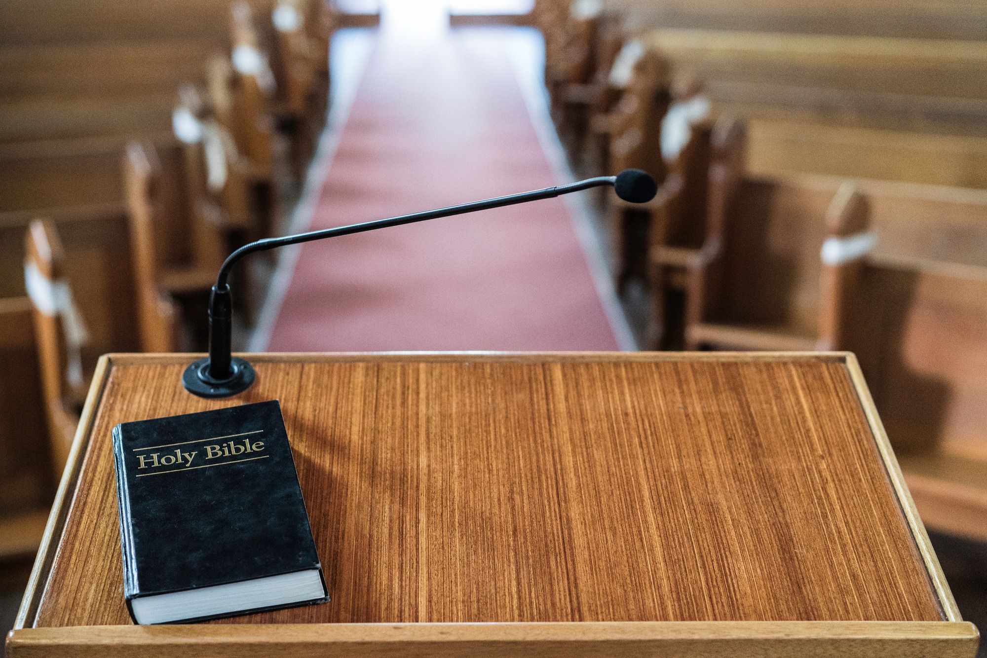A podium with a microphone holds a closed Holy Bible, positioned in a church aisle lined with wooden pews, suggesting a setting for worship or a sermon.