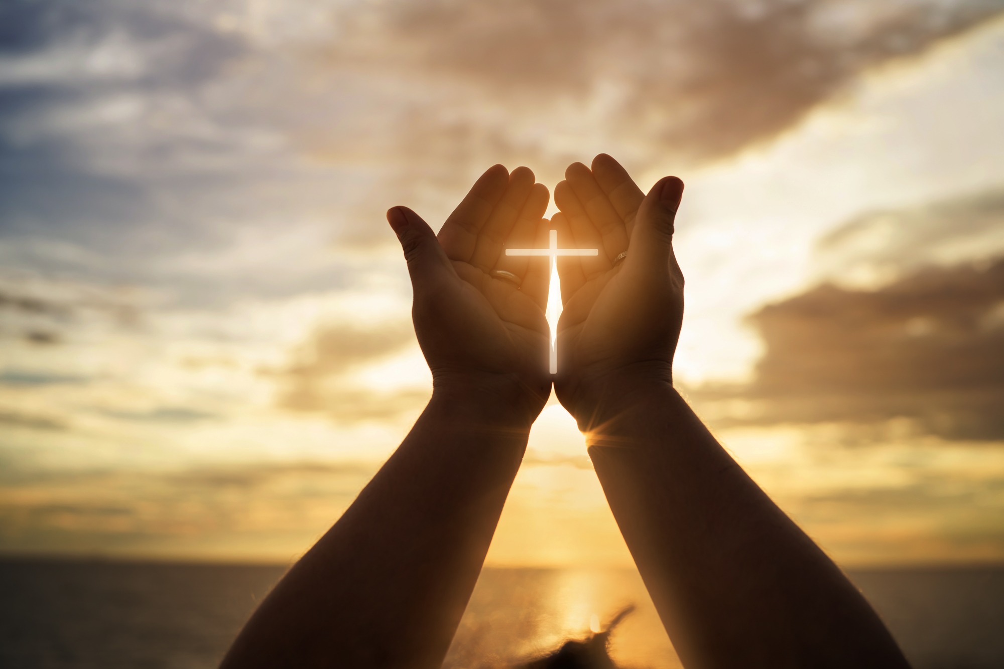 Hands raised against a sunset sky, holding a glowing cross symbolizing faith and hope. The image evokes spirituality and devotion.