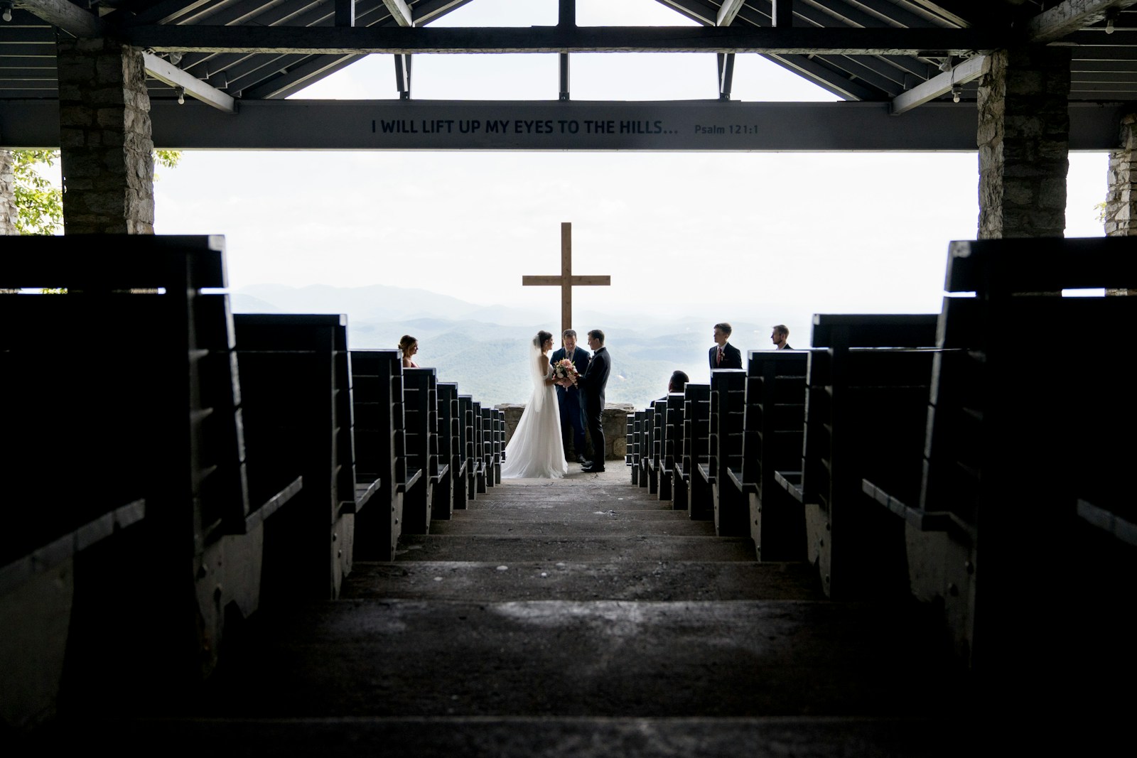 A couple exchanges vows under a cross at an outdoor wedding venue, surrounded by guests and scenic mountains in the background.